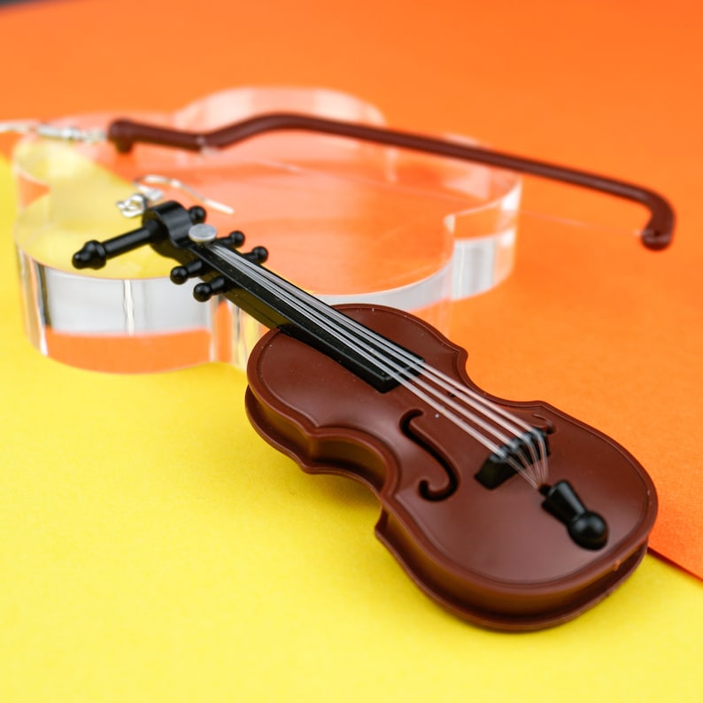 May include: A brown violin-shaped earring with black strings and a silver hook. The earring is on a yellow surface.