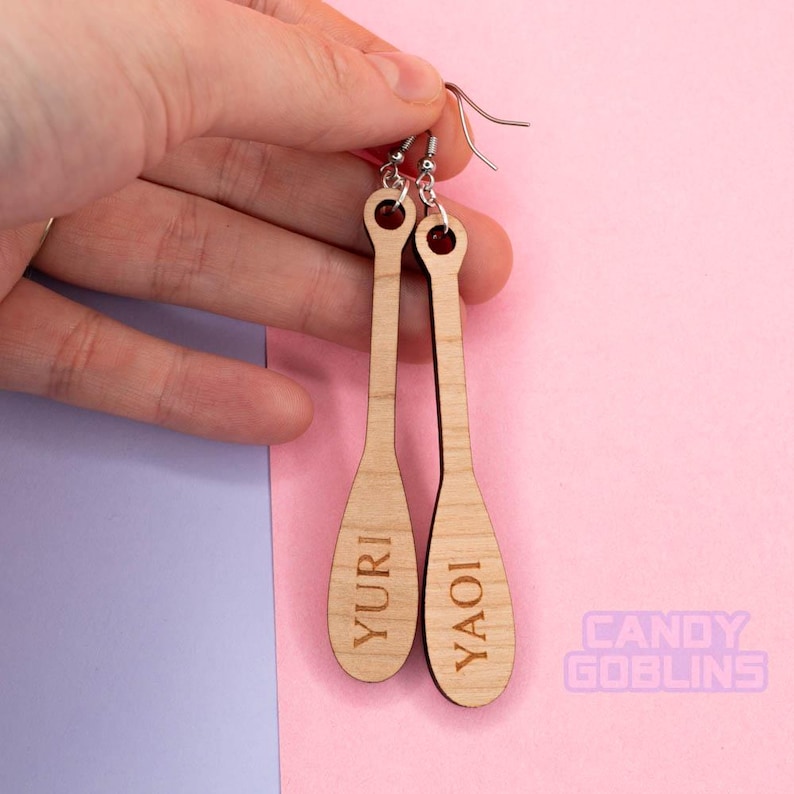 May include: A pair of wooden earrings with the words "YAOI" and "YURI" engraved on them. The earrings are long and slender, with a natural wood finish.