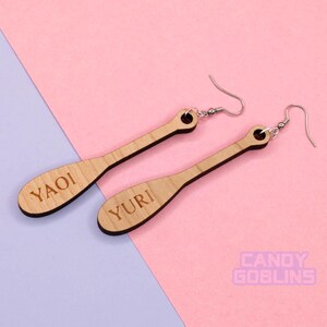 May include: A pair of wooden earrings with silver hooks. The earrings are shaped like paddles and have the words "YAOI" and "YURI" engraved on them.