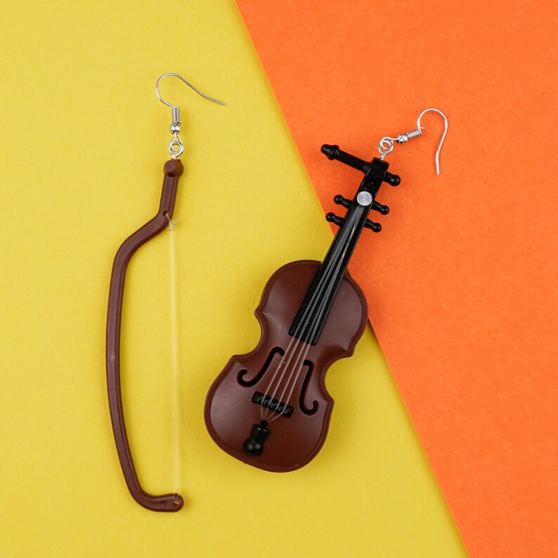 May include: A pair of earrings featuring a violin and a bow. The violin earring is brown and black with a silver hook. The bow earring is brown with a silver hook.