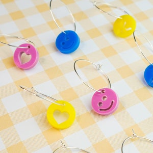 Könnte beinhalten: Ein Set mit sechs silbernen Creolen mit bunten Anhängern. Die Anhänger sind herzförmig und zeigen Smiley-Gesichter. Die Anhänger sind aus Kunststoff und haben eine glänzende Oberfläche.