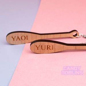 May include: A pair of wooden earrings shaped like baseball bats. The earrings are brown and have the words "YAOI" and "YURI" engraved on them.