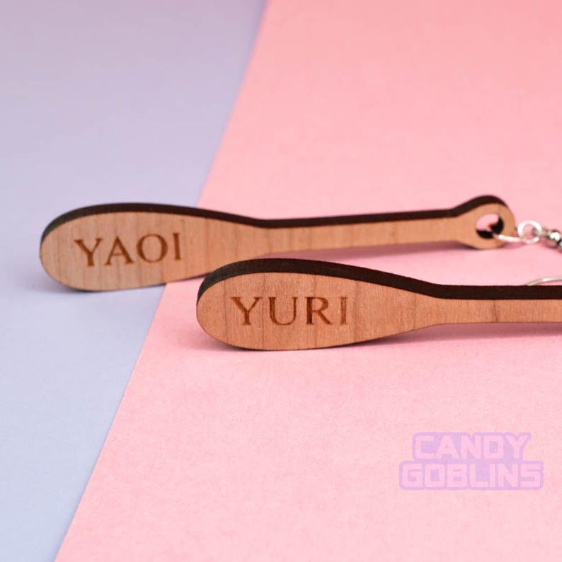 May include: A pair of wooden earrings shaped like baseball bats. The earrings are brown and have the words "YAOI" and "YURI" engraved on them.