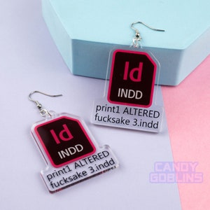 May include: Clear acrylic earrings with a graphic design featuring the Adobe InDesign logo in red and black. The text "print1 ALTERED fucksake 3.indd" is printed below the logo. The earrings have silver-colored fish hook ear wires.