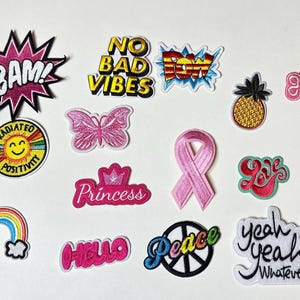 Peut inclure: Un assortiment d'écussons brodés colorés avec divers motifs et textes. Les motifs incluent un arc-en-ciel, un ananas, un papillon et des phrases comme "BAM!", "No Bad Vibes", "Princess" et "Yeah Yeah Whatever!"