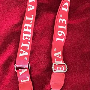 Puede incluir: Correa de hombro roja con texto blanco "MA THETA 1913 DE". La correa tiene herrajes met&aacute;licos plateados y est&aacute; dise&ntilde;ada para sujetarse a un bolso o cartera. El fondo es una tela roja texturizada.