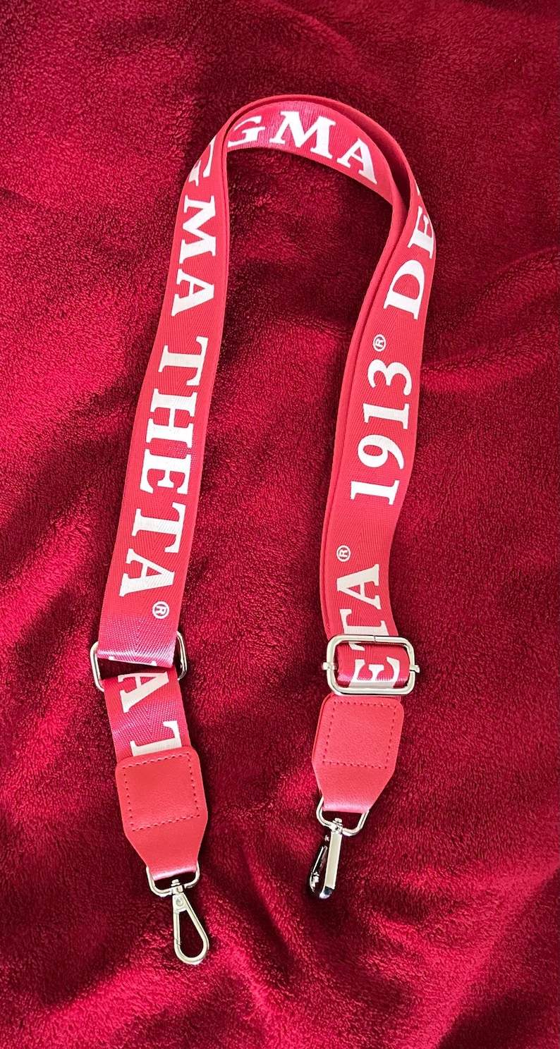 Puede incluir: Correa de hombro roja con texto blanco "MA THETA 1913 DE". La correa tiene herrajes met&aacute;licos plateados y est&aacute; dise&ntilde;ada para sujetarse a un bolso o cartera. El fondo es una tela roja texturizada.
