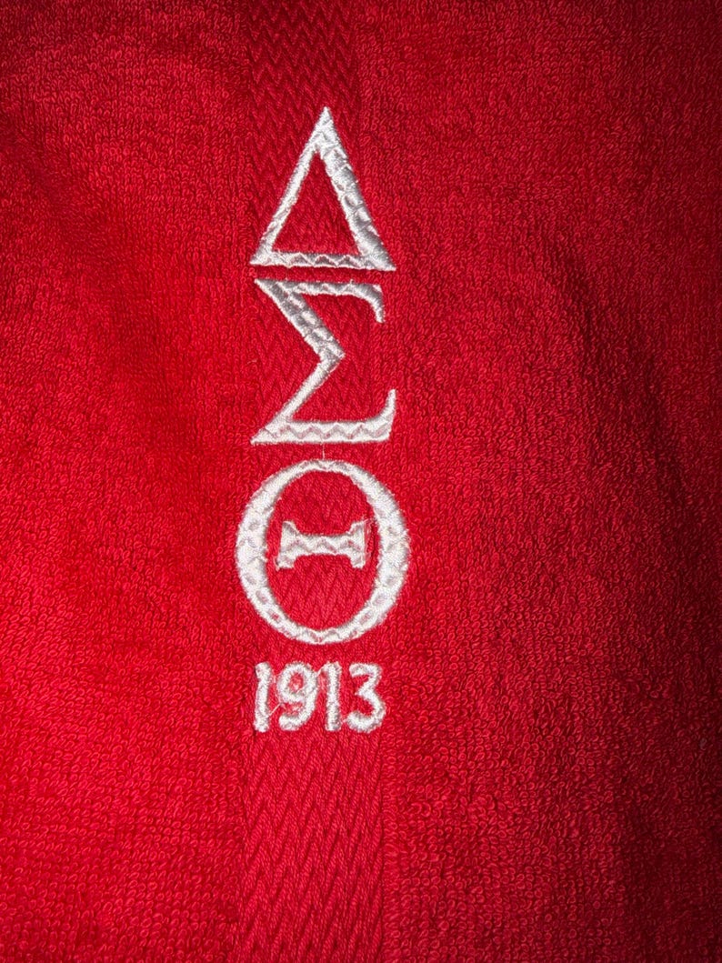 May include: Close-up of a red towel with white embroidered Greek letters and the number 1913. The Greek letters are arranged vertically, with a triangle at the top, followed by a sigma, a zeta, and a circle with a horizontal line through it.