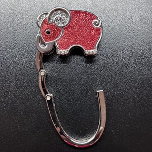 May include: A silver-toned purse hook with a red glitter elephant design. The elephant has rhinestone accents and a silver outline. The hook is designed to hang from a table and hold a purse or bag.