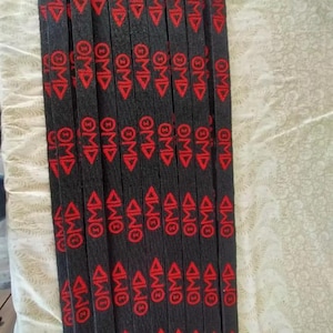 Puede incluir: Un conjunto de cordones negros con texto rojo y un dise&ntilde;o de tri&aacute;ngulo. Cada cord&oacute;n tiene las letras "OMD" repetidas varias veces. Los cordones tienen clips met&aacute;licos plateados en la parte superior.