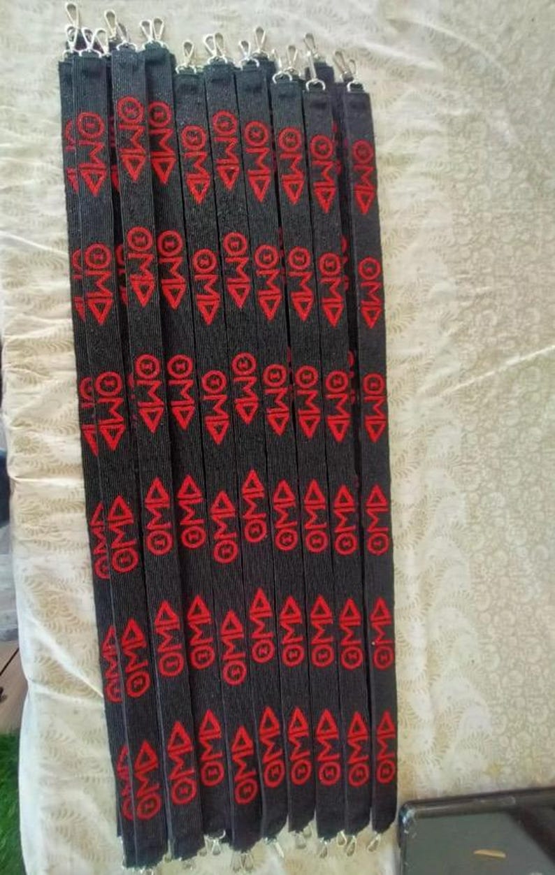 Puede incluir: Un conjunto de cordones negros con texto rojo y un dise&ntilde;o de tri&aacute;ngulo. Cada cord&oacute;n tiene las letras "OMD" repetidas varias veces. Los cordones tienen clips met&aacute;licos plateados en la parte superior.