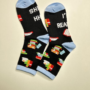 May include: A pair of black socks with light blue cuffs and toes. The socks feature a pattern of colorful books and the text "SHHH I'M READING". The socks are designed for comfort and style, perfect for book lovers.