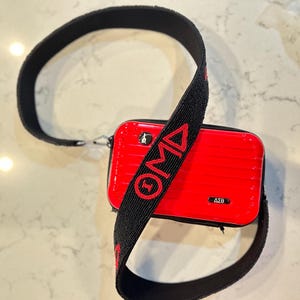 Puede incluir: Un bolso bandolera rectangular rojo con una correa negra. La correa tiene las letras "OMA" en rojo. El bolso tiene un peque&ntilde;o logotipo y las letras "&Delta;&Sigma;&Theta;" en blanco. El bolso est&aacute; sobre una superficie blanca y gris.