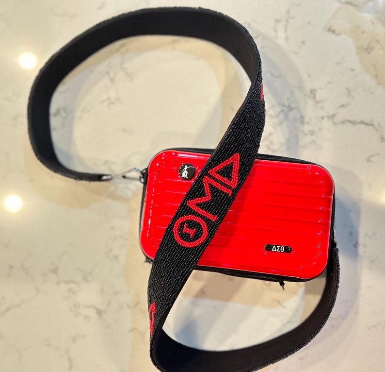 Puede incluir: Un bolso bandolera rectangular rojo con una correa negra. La correa tiene las letras "OMA" en rojo. El bolso tiene un peque&ntilde;o logotipo y las letras "&Delta;&Sigma;&Theta;" en blanco. El bolso est&aacute; sobre una superficie blanca y gris.