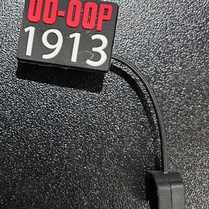 May include: A black and red plastic tag with the text "00-00P 1913" on it. The tag is attached to a black cord with a black plastic loop at the end.