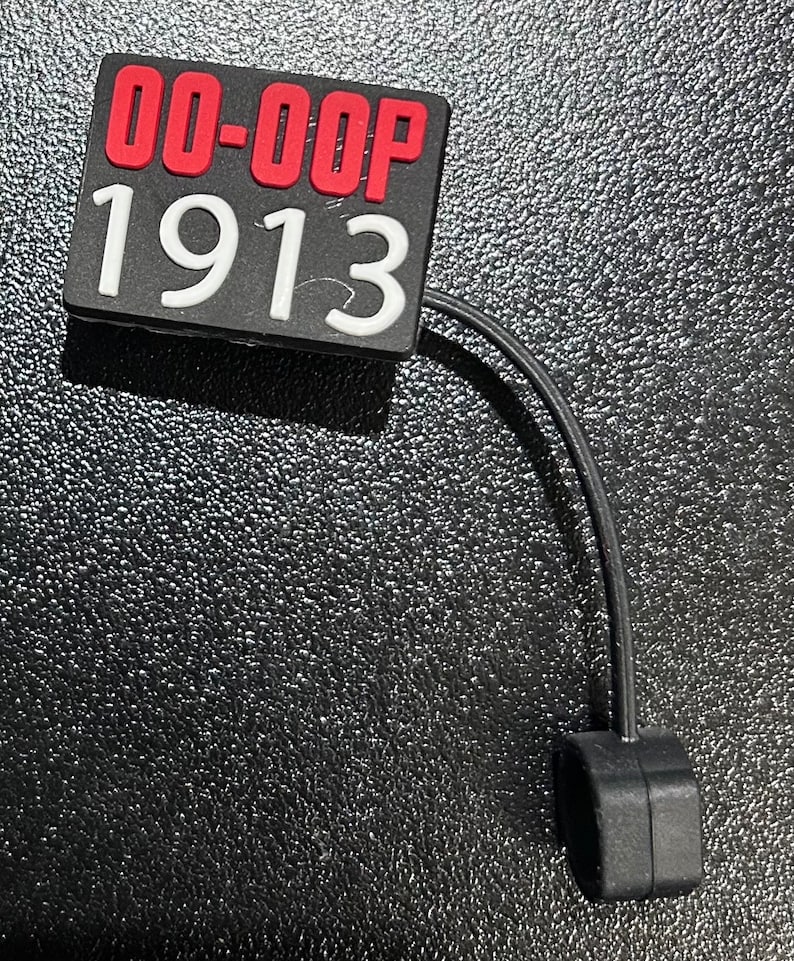 May include: A black and red plastic tag with the text "00-00P 1913" on it. The tag is attached to a black cord with a black plastic loop at the end.