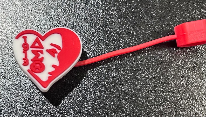 May include: A red heart-shaped charm with a white silhouette of a face and the letters "1913" and "&Delta;&Sigma;&Theta;" inside. The charm is attached to a red string.