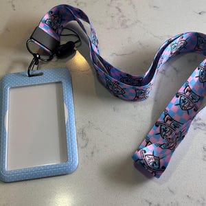 May include: A blue and pink lanyard with a white polka dot pattern and a clear plastic ID card holder. The lanyard features a repeated pattern of cartoon characters with shields.