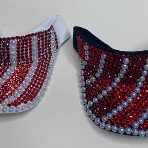 May include: Two visors, one white and one black, are adorned with red and white rhinestones and light blue pearls. The visors are likely for sun protection and fashion, with a decorative design.