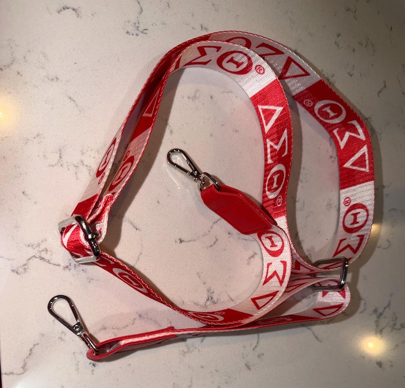 Puede incluir: Un cord&oacute;n rojo y blanco con las letras griegas Sigma, Theta e Iota impresas. El cord&oacute;n tiene un cierre met&aacute;lico y una etiqueta roja. El dise&ntilde;o se repite a lo largo del cord&oacute;n.