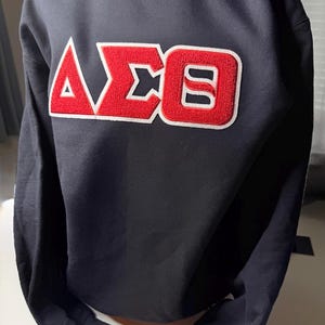 Może przedstawiać: Czarna bluza z haftowanym wzorem greckich liter w kolorze czerwonym i białym. Litery są obrysowane na biało i mają teksturowany wygląd. Bluza ma długie rękawy i okrągły dekolt.