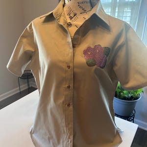 May include: A tan short-sleeved button-down shirt with a pink and green flower design on the left side of the chest.