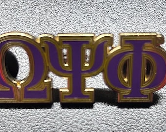Op de afbeelding: Een goudkleurige pin met de Griekse letters Omega, Psi en Phi in paars. De letters hebben een gouden omtrek en staan tegen een getextureerde grijze achtergrond. De pin is waarschijnlijk een broederschaps- of zusterschapsinsigne.