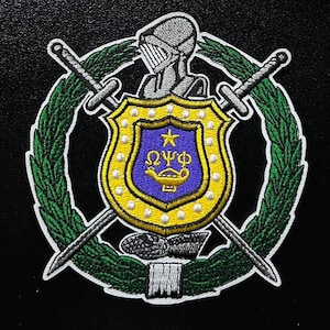 Può includere: Toppa ricamata con uno scudo con una stella e lettere greche, circondato da una ghirlanda verde e spade incrociate. Un elmo da cavaliere è in cima. Lo scudo è giallo e blu.