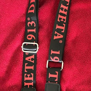 Puede incluir: Correa negra para bolso de hombro con texto rojo "DELTA 1913" y "THETA 1919". La correa tiene herrajes met&aacute;licos plateados y extremos negros de imitaci&oacute;n de cuero. La correa se muestra sobre un fondo rojo.