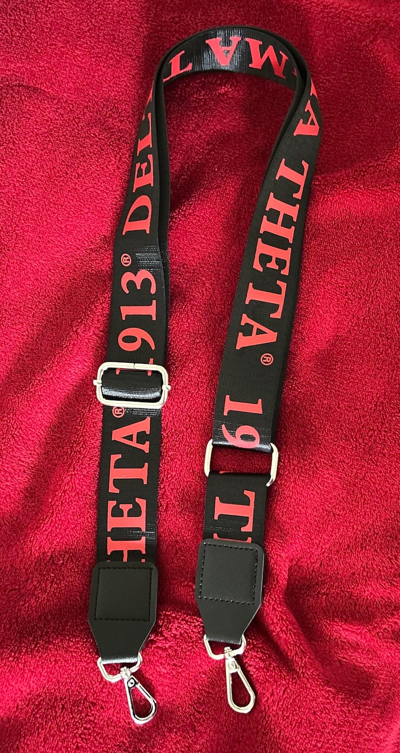 Puede incluir: Correa negra para bolso de hombro con texto rojo "DELTA 1913" y "THETA 1919". La correa tiene herrajes met&aacute;licos plateados y extremos negros de imitaci&oacute;n de cuero. La correa se muestra sobre un fondo rojo.