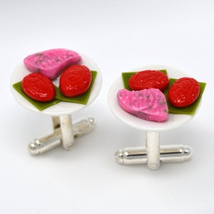 May include: A pair of cufflinks featuring miniature plates with pink and red pastries on top of green leaves. The cufflinks are made of silver.