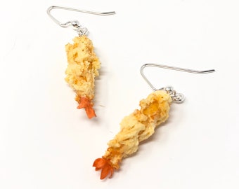 Puede incluir: Un par de pendientes de plata con forma de gambas fritas. Las gambas son de color marrón claro y tienen una textura crujiente. Los pendientes son una forma divertida y única de añadir un toque de fantasía a cualquier atuendo.