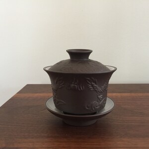 May include: A dark brown ceramic tea pot with a lid and a matching saucer. The pot has a dragon design.