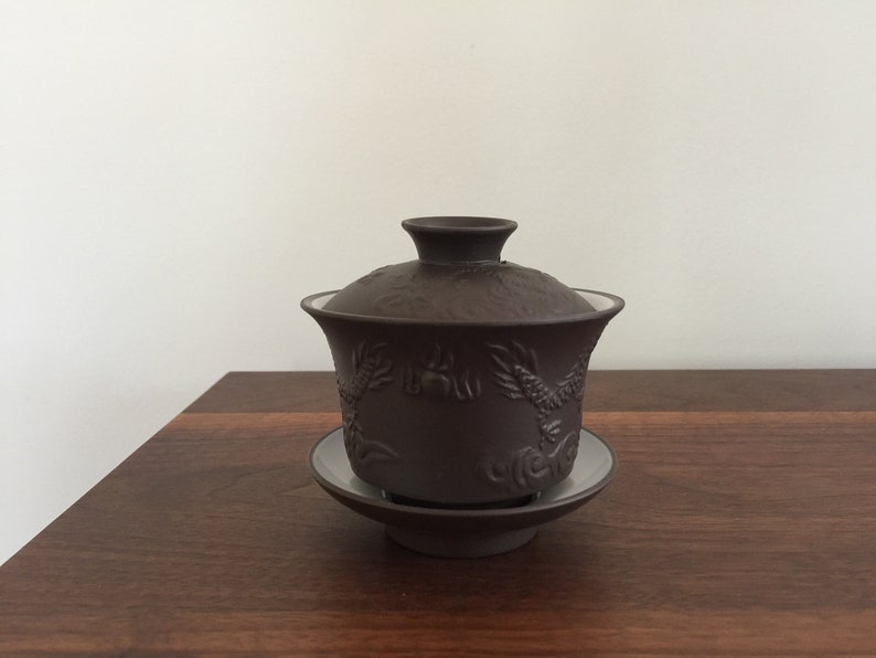 May include: A dark brown ceramic tea pot with a lid and a matching saucer. The pot has a dragon design.