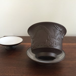 May include: A brown ceramic teacup with a dragon design and a matching saucer. The teacup is sitting on a wooden surface.