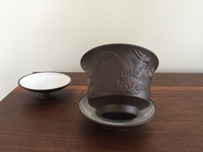 May include: A brown ceramic teacup with a dragon design and a matching saucer. The teacup is sitting on a wooden surface.