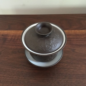 May include: A brown ceramic teacup and lid with a white rim and a dragon design. The teacup is sitting on a saucer.