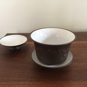May include: A brown ceramic teacup and saucer set with a white interior and intricate embossed designs. The saucer is slightly larger than the teacup.
