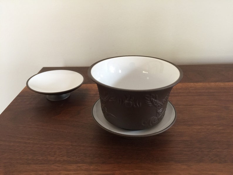 May include: A brown ceramic teacup and saucer set with a white interior and intricate embossed designs. The saucer is slightly larger than the teacup.