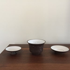 May include: Three ceramic teacups, one with a raised design, on a wooden surface. The cups are brown with white interiors.