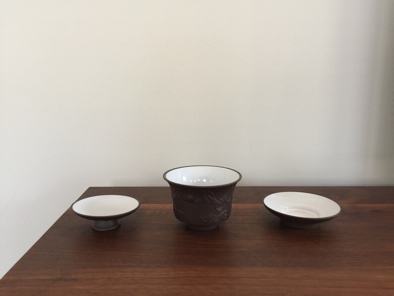 May include: Three ceramic teacups, one with a raised design, on a wooden surface. The cups are brown with white interiors.