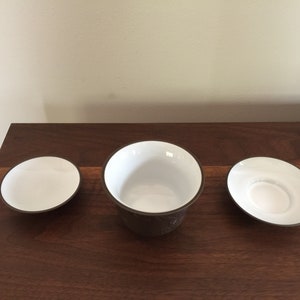 May include: A set of three white ceramic teacups with brown rims. The cups are arranged on a wooden surface.