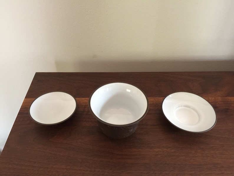 May include: A set of three white ceramic teacups with brown rims. The cups are arranged on a wooden surface.