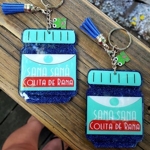 May include: Two blue and green glitter keychains shaped like medicine bottles with a frog charm and a blue tassel. The bottles say "Sana Sana Colita de Rana".