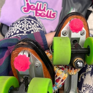 May include: Roller skates with black boots, vivid green wheels, and pink toe stops. A lilac sweatshirt with the text "Jelli Bells" is in the background. The skates are on patterned fabric.