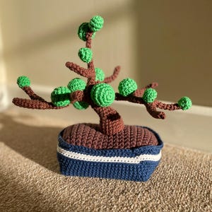 May include: A crocheted bonsai tree with green leaves and a brown pot with a blue and white trim.