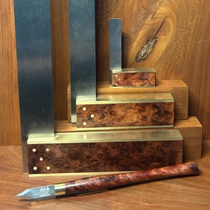 May include: A collection of woodworking tools, including metal squares and a small knife, arranged on a wooden surface. The squares are various sizes, with a polished metal finish. The knife has a wooden handle and a silver blade.