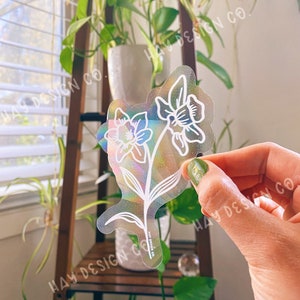 May include: A clear iridescent sticker with a white outline of two daffodils. The sticker has the text "HAY DESIGN CO." printed on the bottom.