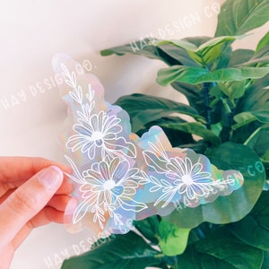 May include: A clear iridescent sticker with a white floral design. The sticker features a cluster of daisies and leaves. The sticker has a holographic finish and is perfect for adding a touch of whimsy to any project.