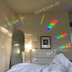 Puede incluir: Un dormitorio con una c&oacute;moda blanca, un espejo dorado y una cama blanca con ropa de cama blanca. Las paredes son blancas y hay reflejos de prisma arco&iacute;ris en las paredes. El texto "HAY DESIGN CO." est&aacute; escrito en las paredes.